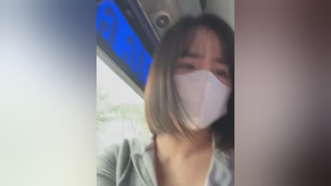 Media: A video of an Asian woman with shoulder-length brown hair and a surgical mask, wearing a light green cardigan, seated in a car with a blurred background featuring a blue sign.