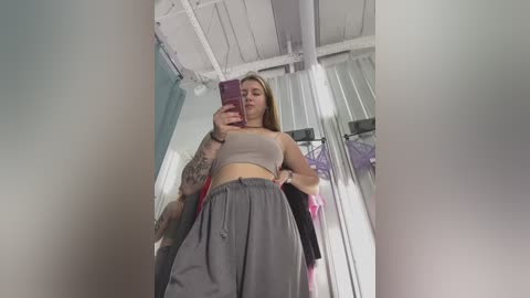 Media: Video of a young woman with long brown hair, light skin, and medium build, wearing a gray crop top and high-waisted pants, taking a selfie in a modern, white-walled room with large windows.