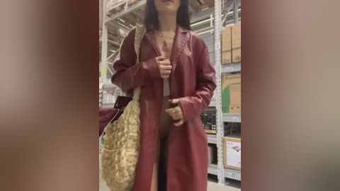 Media: Video of a woman in a red trench coat carrying a beige handbag, standing in a warehouse with shelving and boxes in the background.