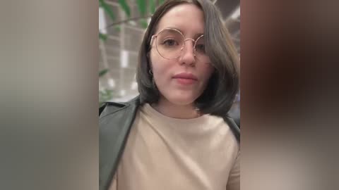 Media: Video of a fair-skinned woman with straight, shoulder-length brown hair, wearing clear-framed glasses and a beige top. She has a neutral expression. Background shows blurred green plants and white walls.