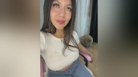 Media: Video of a Latina woman with long, dark hair, wearing a white ribbed crop top and high-waisted blue jeans, seated on a pink chair. Background features a white wall and a black curtain, with a small dog visible on the floor.