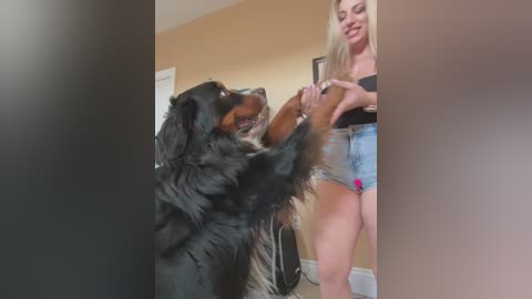 Video of a woman in a black top and denim shorts standing with a large, black-and-brown dog, both smiling. Background shows beige walls and a white door.