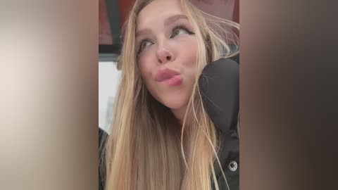 A video of a young blonde woman with long hair, wearing a black jacket, making a pouty face, blurred background.