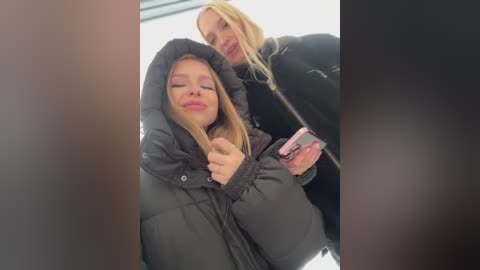 Media: Video of two young women with blonde hair, one wearing a black puffer jacket, the other a black coat, smiling, holding pink phone, blurred background.