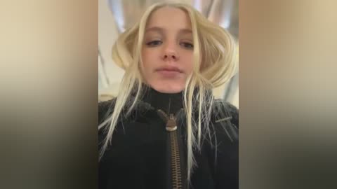 Media: Video of a blonde woman with long, tousled hair, wearing a black jacket with a fur-lined hood, standing against a blurred background.