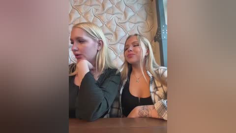 Media: Video of two young blonde women, one with shoulder-length hair and a green top, the other with long hair and a checkered shirt, sitting side by side in a dimly lit restaurant.