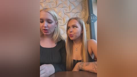 Media: Video of two young women, one blonde and one brunette, sitting at a table in a dimly lit restaurant. They have a playful, pouty expression. The background shows a patterned wall and a lamp.