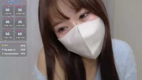 Media: Video of an Asian woman with long, straight brown hair, wearing a white surgical mask, light blue off-shoulder top, and a digital screen showing weather forecast with temperatures and humidity levels.