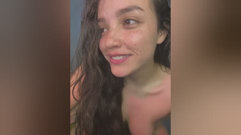 Media: Video of a young woman with wet, dark, curly hair, fair skin, and freckles, smiling slightly, in a blurred indoor setting.