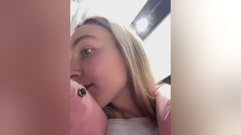 Video of a young woman with light skin and long blonde hair, wearing a pink shirt, lying on her side, looking at a pink stuffed animal with a black button eye. Background includes a white ceiling and a spotlight.