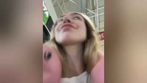 A candid video of a smiling, light-skinned woman with straight blonde hair, wearing a white tank top, captured from a low angle, emphasizing her upward gaze.