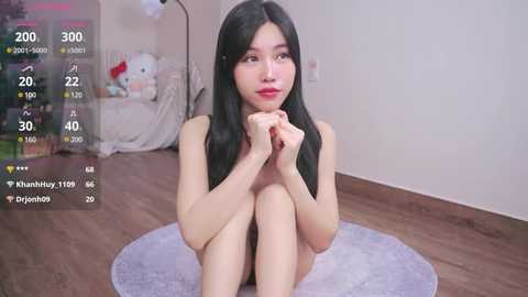 Media: A video of an East Asian woman with long black hair, seated nude on a plush rug, gazing thoughtfully. The room features a soft bed and a lamp in the background.