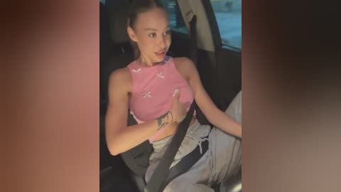 Media: Video of a light-skinned, slim woman with long brown hair tied back, wearing a pink crop top and grey pants, seated in a car with seat belt on.