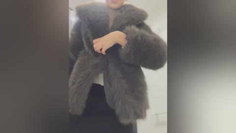 Media: Video of a person wearing a luxurious, dark gray, fur-trimmed coat over a white top and black pants, standing indoors with a blurred, neutral background.