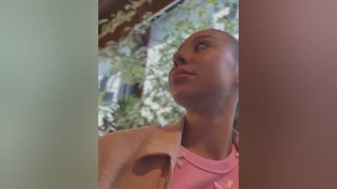 Video of a young woman with light skin, wearing a pink t-shirt, looking upward through a window at a lush, green garden outside.