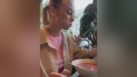 A video of a woman with blonde hair tied back, wearing a beige jacket over a pink shirt, eating soup at a table, surrounded by lush greenery outside.