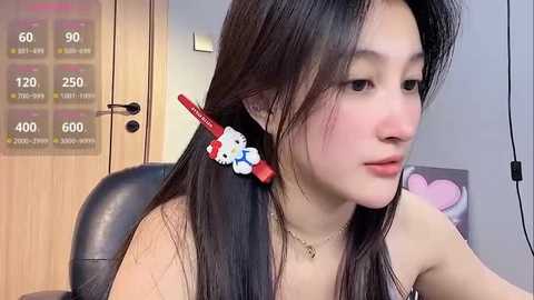 Media: Video of an East Asian woman with long black hair, adorned with a red Hello Kitty hair clip, wearing a gold necklace, sitting indoors.