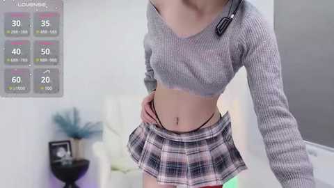 Media: Video of a slender, light-skinned woman with a flat stomach, wearing a cropped ribbed sweater and a short plaid skirt, standing in a minimalist, white room with a fitness tracker display on the wall.