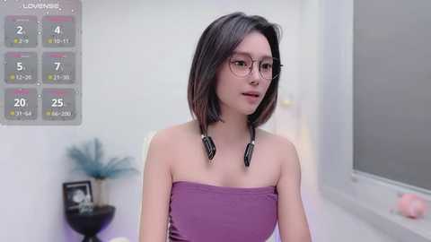 Media: Video of a young woman with a slender physique, light skin, and shoulder-length dark hair. She wears a strapless purple dress and large round glasses, standing in a modern, minimalistic room with a calendar and a small potted plant in the background.