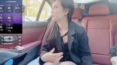 Media: Video of a young woman with long dark hair, wearing a black blazer and a revealing black lace top, sitting in a car with red leather seats.