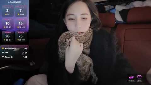 Media: Video of a young woman with long black hair, wearing a black coat, holding a brown fur scarf, sitting in a car, with a live streaming app interface on the left.