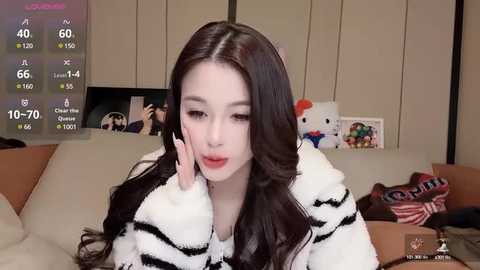 Media: Video of a young Asian woman with long black hair, fair skin, and makeup, wearing a white and black striped sweater, sitting in a cozy living room with plush toys and a TV displaying weather information.