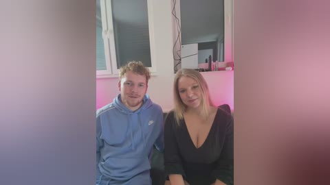 Media: Video of a young Caucasian couple with fair skin, the man in a blue jacket and the woman in a low-cut black top, sitting indoors.