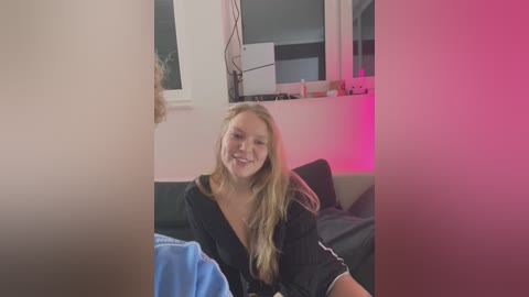 Media: A video of a blonde woman in a black robe, smiling, sitting on a bed with pink lighting in the background.