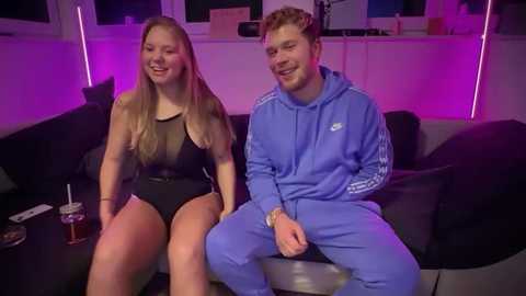 Media: Video of a young, smiling couple in casual clothing, sitting on a black couch in a dimly-lit room with purple lighting.