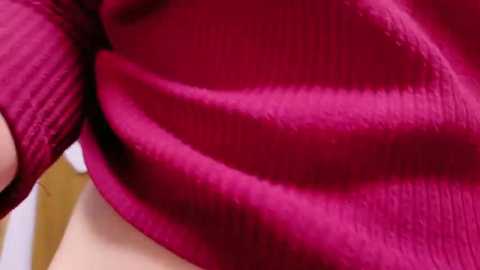 Media: Close-up video of a person's hand lifting a vibrant, deep magenta sweater with a ribbed texture, revealing the smooth, light skin underneath. The background is blurred, emphasizing the rich color and soft fabric of the garment.