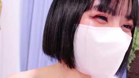 Media: A close-up video of an East Asian woman with a short black bob haircut, wearing a white face mask, with a blurred background of a light blue curtain and a potted plant.