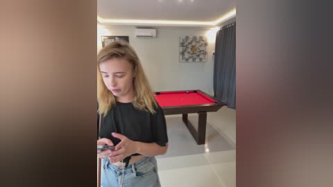 Media: Video of a blonde woman in a black top and jeans playing pool in a modern, brightly-lit room with a red pool table, framed art, and dark curtains.