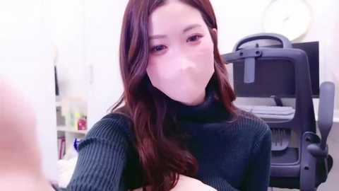 Media: Video of a young Asian woman with long, dark hair, wearing a black turtleneck sweater and a surgical mask, sitting in an office chair with a desk behind her.