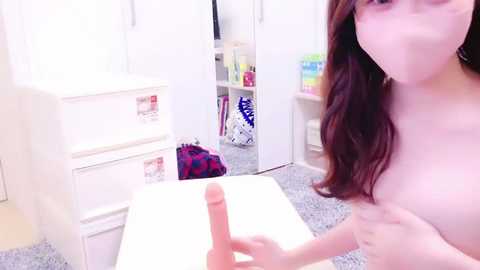 Media: Video of a nude woman with long brown hair and a face mask, holding a large, realistic-looking dildo in a minimalist, bright room with white furniture, colorful storage bins, and a fluffy carpet.