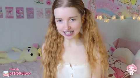 Media: A video of a young, fair-skinned woman with long, curly, blonde pigtails, wearing a white, strapless top. The background features pastel-colored decor, including plush toys and fairy lights. She has a friendly smile.