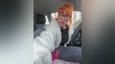 Video of a young woman with orange hair, wearing black glasses and a white fluffy blanket, sleeping in a car seat.