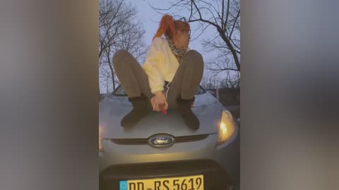 Media: Video of a woman in a yellow jacket, brown pants, and black boots, sitting on a silver car's hood, with a bare tree background and a license plate reading \"PNS 5419.\