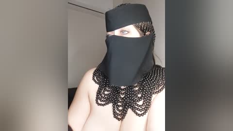 Media: Video of a light-skinned woman wearing a black veil and elaborate beaded necklace, partially exposing her bare breasts. The background is plain and neutral-colored, possibly a room with minimal decoration.