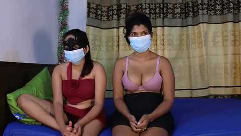 Media: Video of two women in face masks, sitting on a bed with blue sheets, wearing red and pink lingerie; background includes a curtain with floral patterns.