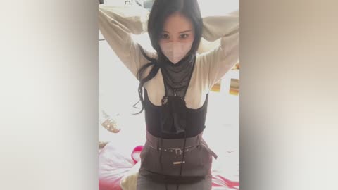 Media: Video of an Asian woman with long black hair, wearing a beige top, black vest, and brown pants, arms raised, indoors, blurry background.