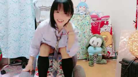 Media: Video of a young Asian woman with short black hair, wearing a plaid shirt and black knee-high socks, kneeling on a colorful rug, smiling in a playful, cluttered room with plush toys, a chair, and festive decorations.