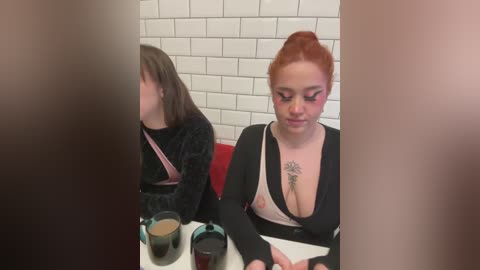 Media: Video of two women in a diner: one with auburn hair, black top, large breasts, and tattoo; the other with shoulder-length brown hair, black velvet top, and pink strap. White tiled wall background.