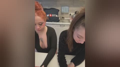 Media: Video of two fair-skinned women with red hair, one topless with a tattoo, and the other in a black turtleneck, sitting at a kitchen table.