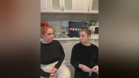 Media: Video of two women in a kitchen: a red-haired woman in black, and a brunette in black, both with surprised expressions, in a modern white cabinetry setting.