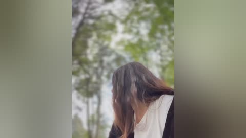 Media: Video of a person with long, straight brown hair, wearing a white tank top, seen from behind, standing in a forest with blurred green trees and a light sky background.