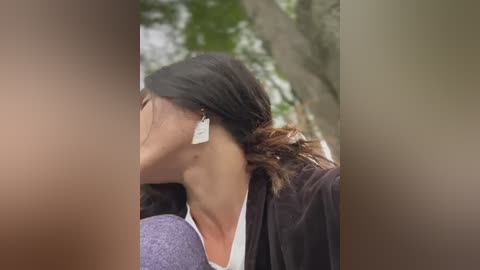 Media: A video of a woman with dark hair, wearing a white shirt and a black jacket, kissing another person whose face is blurred. The background features trees and a blurry sky.