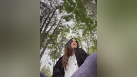 Media: Video of a young woman with long, dark hair, wearing a white top and black jacket, seated outdoors against a background of lush green trees.