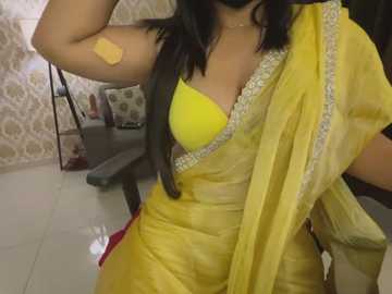 Media: Video of a woman with medium skin tone, wearing a yellow sari with a yellow bra peeking through, standing indoors with floral wallpaper and a grey table in the background.