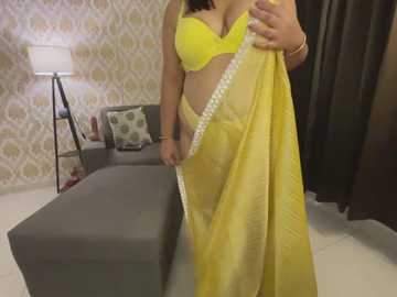 Media: Video of a woman in a bright yellow saree with a matching bra, standing in a modern living room with a grey couch, patterned wallpaper, and a black curtain.