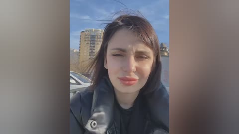 Media: Video of a young woman with straight dark hair, wearing a black coat, squinting in the sunlight. Background features a cityscape with a high-rise building and parked cars.